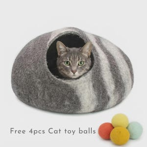 May include: A dome-shaped gray and white felted wool cat cave with a circular opening containing a tabby cat. Four small, round, solid-colored felted wool balls in yellow, orange, and light blue are positioned below with the text Free 4pcs Cat toy balls.