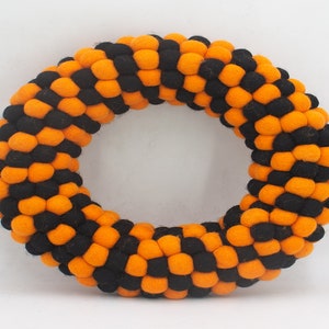 Puede incluir: Una corona de Halloween hecha de bolas de fieltro naranja y negro. La corona es circular y tiene un di&aacute;metro de aproximadamente 30 cm.