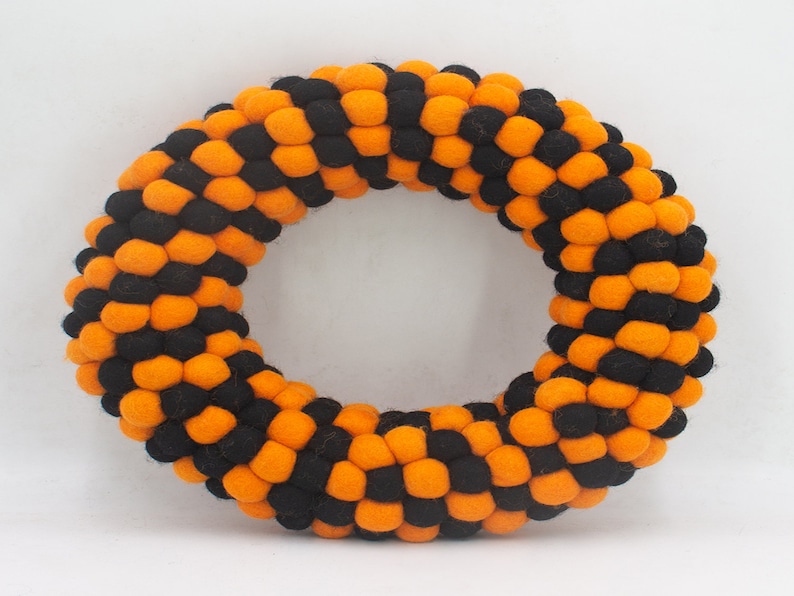 Puede incluir: Una corona de Halloween hecha de bolas de fieltro naranja y negro. La corona es circular y tiene un di&aacute;metro de aproximadamente 30 cm.