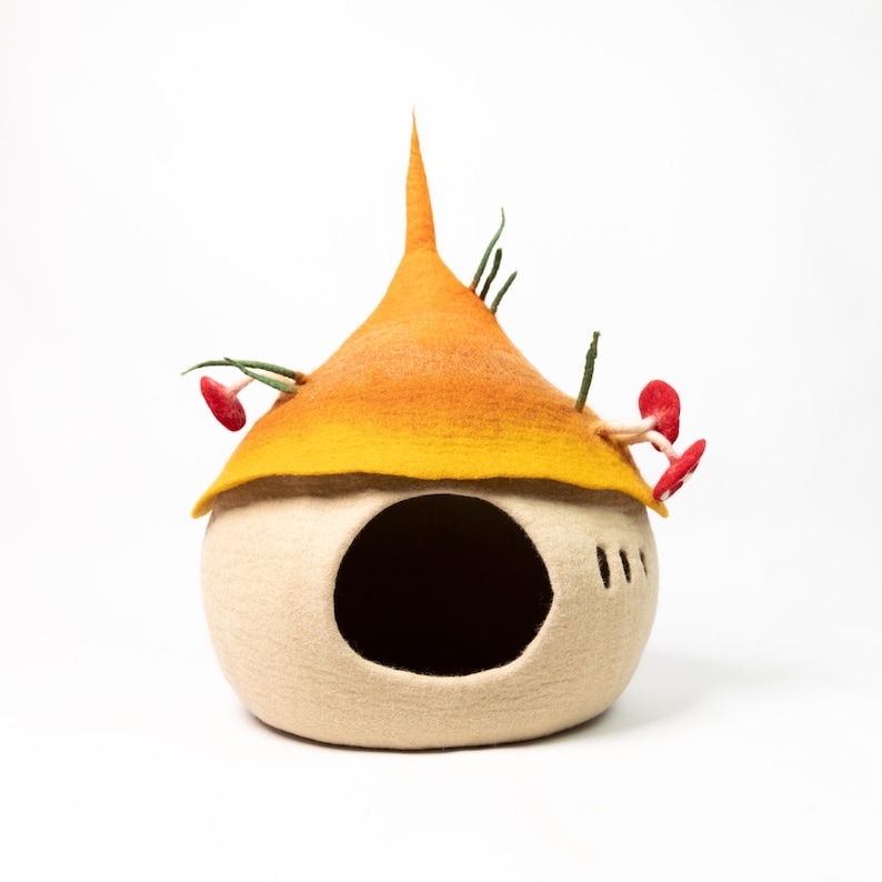 May include: A felted wool pet cave featuring a cream-colored rounded base with a large circular opening and three small vertical slits. The top is a pointed orange and yellow roof adorned with small red mushroom accents and green stems.
