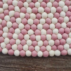 May include: A round pink and white felt rug made of small balls of wool. The rug is on a wooden surface.