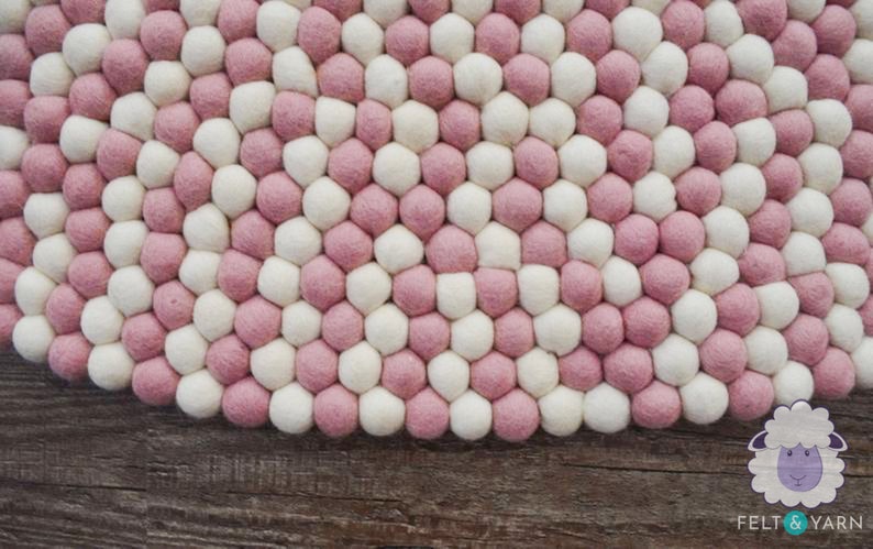 May include: A round pink and white felt rug made of small balls of wool. The rug is on a wooden surface.