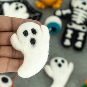 May include: A white felt ghost with black eyes and a mouth, held in a hand. The ghost is a Halloween decoration.