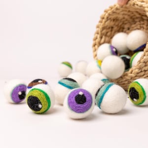 May include: A basket overflowing with felt eyeball ornaments. The eyeballs are white with various colored irises and pupils. The basket is made of woven straw.