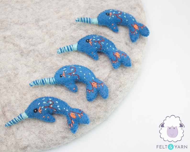 May include: Four blue felt narwhal ornaments with embroidered details. Each narwhal has a light blue, curled horn and a black eye. The narwhals are arranged in a row on a light gray surface.