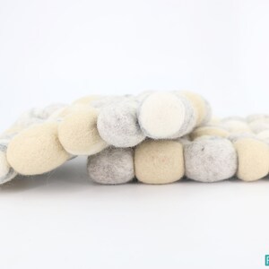 Puede incluir: Un salvamanteles de fieltro hecho a mano en una paleta de colores neutros. El salvamanteles está compuesto por múltiples bolas de fieltro en tonos crema, gris y beige. Las bolas de fieltro están dispuestas en un patrón similar a una cuadrícula, creando una superficie texturizada.