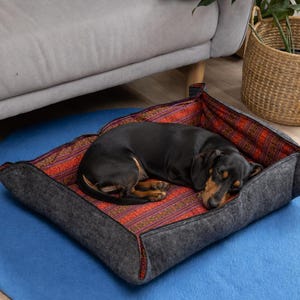 May include: A black and tan dachshund sleeping in a square pet bed. The bed features a dark gray felt exterior and an interior lining with a vibrant orange, red, and black geometric pattern. The sides of the bed are folded up to create a cozy enclosure.