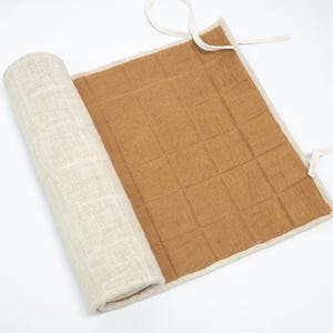 May include: A rolled-up beige linen fabric next to a flat, open brown fabric with a grid pattern. The edges are trimmed with a lighter beige fabric and have white ties. The item appears to be a mat or wrap.