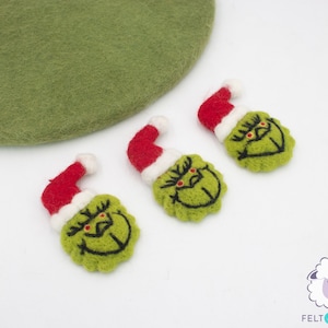 Könnte beinhalten: Drei Filz-Anhänger in Form des Grinchs aus dem Film Wie der Grinch Weihnachten gestohlen hat. Jeder Anhänger ist grün mit einer roten Weihnachtsmütze und schwarzen Gesichtszügen. Die Anhänger befinden sich auf einem weißen Hintergrund.