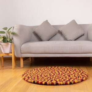 May include: A round, multi-colored felt ball rug with a mix of yellow, orange, red, and purple balls. The rug is in front of a gray sofa with two gray throw pillows.