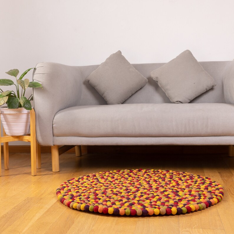 May include: A round, multi-colored felt ball rug with a mix of yellow, orange, red, and purple balls. The rug is in front of a gray sofa with two gray throw pillows.