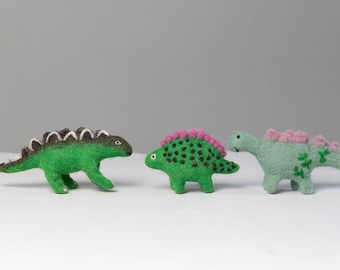May include: Three felt dinosaur toys. One is green with black spots and pink spines, one is green with black stripes and white spines, and one is light green with pink spines and green leaves.