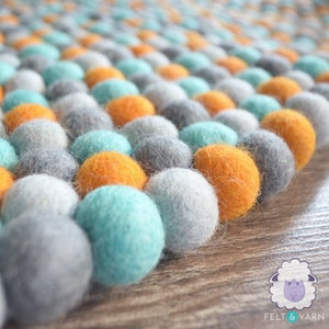 May include: A close-up of a felt ball rug with a pattern of orange, gray, light blue, and white balls. The rug is made of many small, round felt balls arranged in rows. The rug is on a wooden surface.