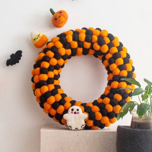 Puede incluir: Una corona de Halloween hecha de bolas de fieltro naranja y negro. La corona est&aacute; decorada con calabazas de fieltro, un fantasma y un ma&iacute;z dulce.