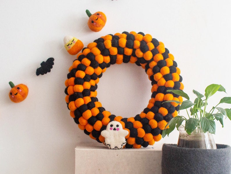 Puede incluir: Una corona de Halloween hecha de bolas de fieltro naranja y negro. La corona est&aacute; decorada con calabazas de fieltro, un fantasma y un ma&iacute;z dulce.