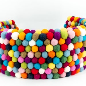 Pu&ograve; includere: Un cuscino a forma di mezzaluna composto da palline di feltro rotonde e multicolori. Il cuscino &egrave; fatto di molte piccole palline di feltro cucite insieme.