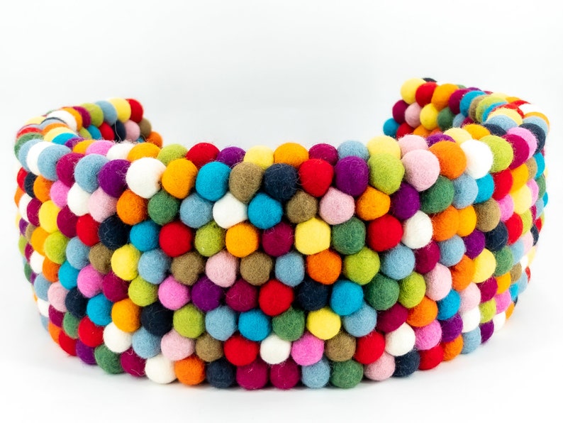Pu&ograve; includere: Un cuscino a forma di mezzaluna composto da palline di feltro rotonde e multicolori. Il cuscino &egrave; fatto di molte piccole palline di feltro cucite insieme.
