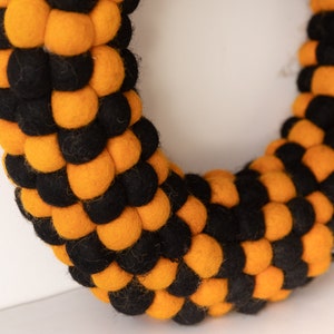Puede incluir: Una corona de bolas de fieltro negra y naranja. La corona est&aacute; hecha de peque&ntilde;as bolas de fieltro redondas que est&aacute;n dispuestas en un patr&oacute;n circular. Las bolas alternan negro y naranja.