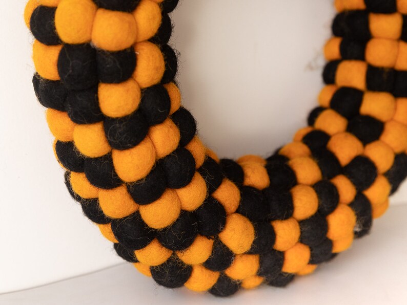 Puede incluir: Una corona de bolas de fieltro negra y naranja. La corona est&aacute; hecha de peque&ntilde;as bolas de fieltro redondas que est&aacute;n dispuestas en un patr&oacute;n circular. Las bolas alternan negro y naranja.