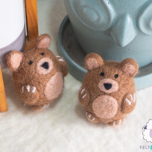Könnte beinhalten: Zwei braune Filz-Bären-Ornamente mit weißen Akzenten. Die Bären haben schwarze Augen und Nasen. Die Bären sitzen auf einer weißen Oberfläche.