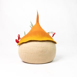 May include: A needle-felted sculpture featuring a round, beige base topped with a pointed, gradient orange and yellow cap. Small, red mushroom-like accents and thin, green, grass-like fibers emerge from the sides of the textured, soft-looking object.
