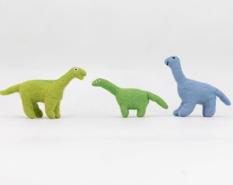 May include: Three felt dinosaur toys. One is green, one is light blue, and one is a darker shade of green. They are all standing on a white surface.