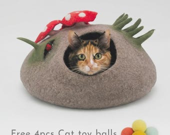May include: A felted gray cat cave shaped like a mound with a red and white spotted mushroom top and green grass-like accents. A calico cat is visible inside the opening. Four small, colorful felt balls are placed in front with the text Free 4pcs Cat toy balls.