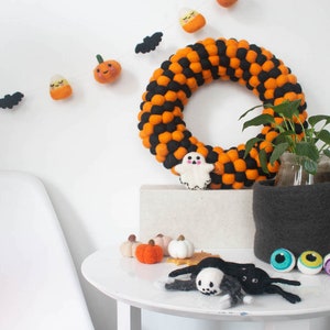 Puede incluir: Una corona de Halloween hecha de bolas de fieltro naranja y negro. La corona est&aacute; decorada con un fantasma de fieltro, calabazas de fieltro y un ma&iacute;z dulce de fieltro. Tambi&eacute;n hay murci&eacute;lagos y calabazas de fieltro colgando de la pared detr&aacute;s de la corona.