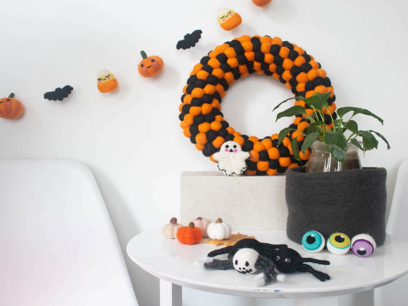 Puede incluir: Una corona de Halloween hecha de bolas de fieltro naranja y negro. La corona est&aacute; decorada con un fantasma de fieltro, calabazas de fieltro y un ma&iacute;z dulce de fieltro. Tambi&eacute;n hay murci&eacute;lagos y calabazas de fieltro colgando de la pared detr&aacute;s de la corona.