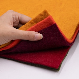May include: A stack of four felt sheets in different colors: mustard yellow, orange, burgundy, and forest green. The sheets are folded and layered, with the top sheet being mustard yellow.