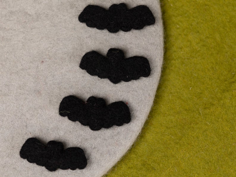 May include: Four black felt bats are arranged in a vertical line on a light gray felt background. The bats are all facing the same direction, with their wings spread out. The background is a light gray felt, and the bats are a dark black felt.