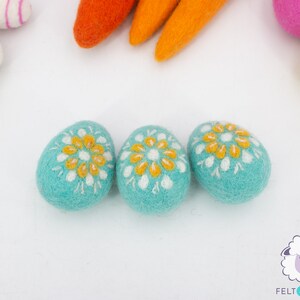 K&ouml;nnte beinhalten: Drei t&uuml;rkisfarbene Filz-Ostereier mit wei&szlig;en und gelben gestickten Blumenmustern.