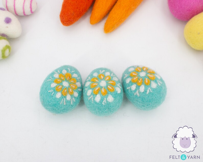 K&ouml;nnte beinhalten: Drei t&uuml;rkisfarbene Filz-Ostereier mit wei&szlig;en und gelben gestickten Blumenmustern.
