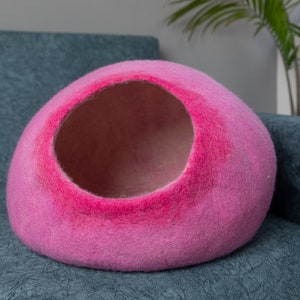 May include: A pink felt cat bed shaped like a dome with a large opening. The bed is sitting on a blue couch.