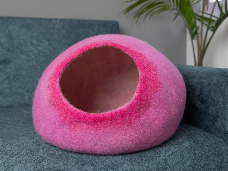 May include: A pink felt cat bed shaped like a dome with a large opening. The bed is sitting on a blue couch.