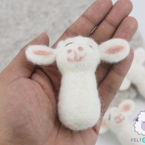 May include: A white felt sheep with pink ears and a smiling face. The sheep is being held in a hand.
