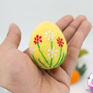 K&ouml;nnte beinhalten: Ein gelbes Filz-Osterei mit gestickten wei&szlig;en und roten Blumen.