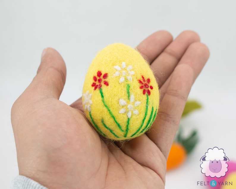 K&ouml;nnte beinhalten: Ein gelbes Filz-Osterei mit gestickten wei&szlig;en und roten Blumen.