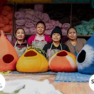May include: Four women stand in front of a wall of colourful yarn, holding handmade felt cat beds in orange, blue, and red. The beds are shaped like teardrops and have a hole for the cat to enter. The women are wearing aprons and smiling. The text "goodweave+ CERTIFIED" and "Sedex AUDITED" is visible in the image.
