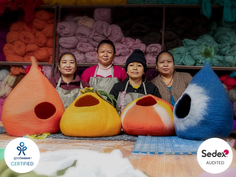 May include: Four women stand in front of a wall of colourful yarn, holding handmade felt cat beds in orange, blue, and red. The beds are shaped like teardrops and have a hole for the cat to enter. The women are wearing aprons and smiling. The text "goodweave+ CERTIFIED" and "Sedex AUDITED" is visible in the image.