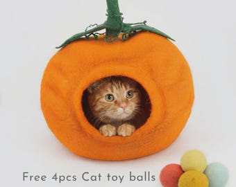 May include: A bright orange felted pumpkin-shaped cat cave with a green stem and leaf top. An orange tabby cat is visible inside the opening. Four small, round, multi-coloured felted wool balls are placed in front of the cat cave.