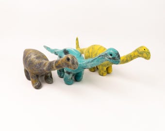 May include: Three felt dinosaur toys. One is gray, one is teal, and one is yellow.