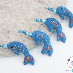 May include: Four blue felt narwhal ornaments with embroidered details. Each narwhal has a light blue spiral horn and a black eye. The narwhals are arranged in a row on a light gray surface.