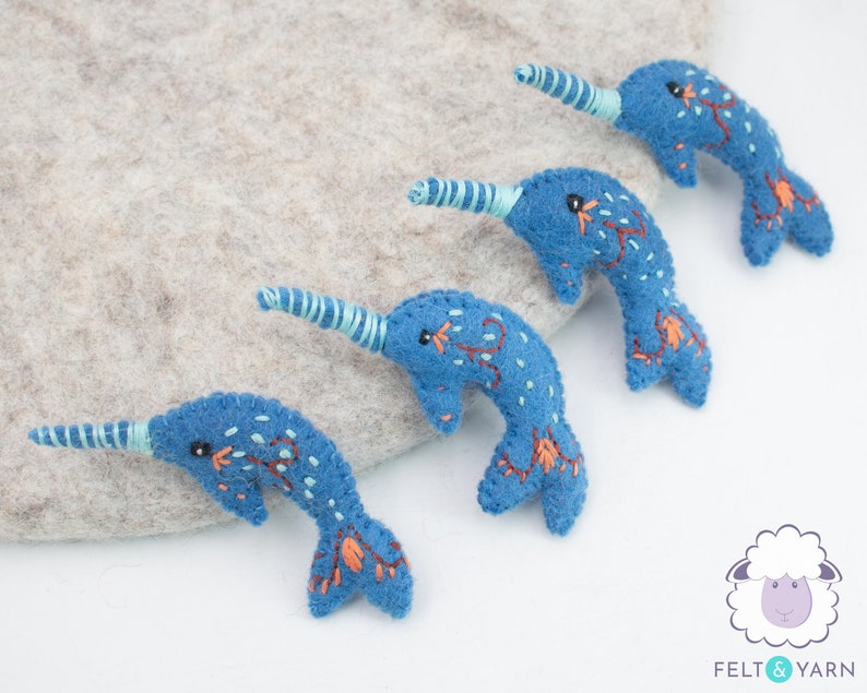 May include: Four blue felt narwhal ornaments with embroidered details. Each narwhal has a light blue spiral horn and a black eye. The narwhals are arranged in a row on a light gray surface.