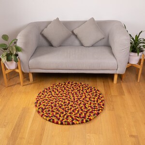 May include: A round, multi-colored felt rug with a variety of shades of red, orange, yellow, and green. The rug is made of small, individual felt balls that are tightly packed together. The rug is sitting on a wooden floor in front of a gray sofa.