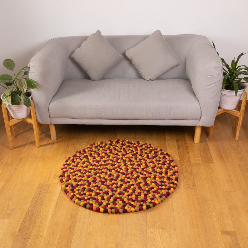 May include: A round, multi-colored felt rug with a variety of shades of red, orange, yellow, and green. The rug is made of small, individual felt balls that are tightly packed together. The rug is sitting on a wooden floor in front of a gray sofa.