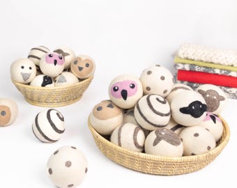 May include: Two woven baskets filled with round, felted wool balls in cream, beige, brown, and black. The balls feature various designs, including animal faces, polka dots, and swirling lines. Some balls are scattered outside the baskets.