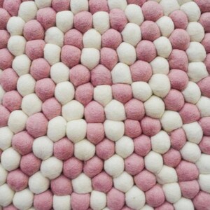 May include: A close-up of a round, pink and white felt ball rug. The balls are arranged in a circular pattern, creating a unique and modern design.