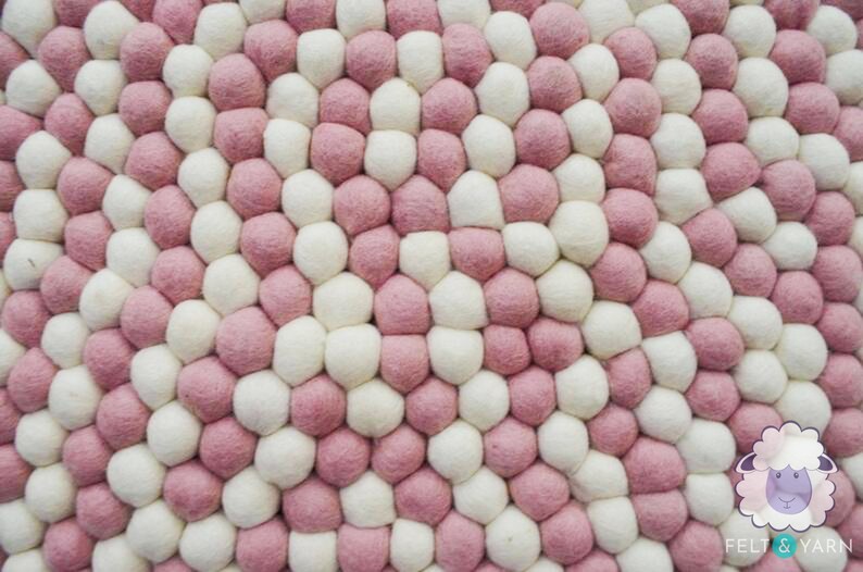 May include: A close-up of a round, pink and white felt ball rug. The balls are arranged in a circular pattern, creating a unique and modern design.