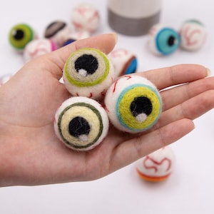 May include: A hand holding three felt eyeball ornaments. The eyeballs are green, yellow, and white with black pupils and red veins.